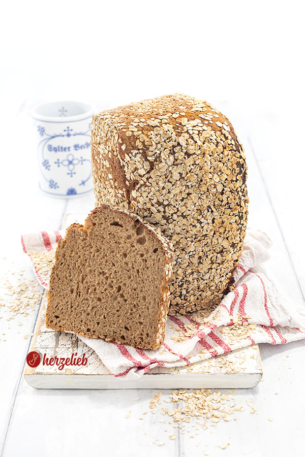 Ein mit Haferflocken bestreutes Brot nach einem Hafer-Dinkel-Kastenbrot Rezept von herzelieb. Der Brotlaib steht auf der Schnittfläche, daran lehnt eine Scheibe davon.