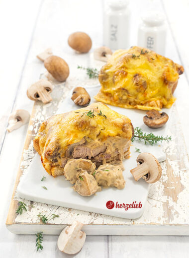 Ein angeschnittener Jägerotast auf einem Porzellanbrett. Dahinter ein weiteres Toast. Dekoriert mit Thymian und Champignons