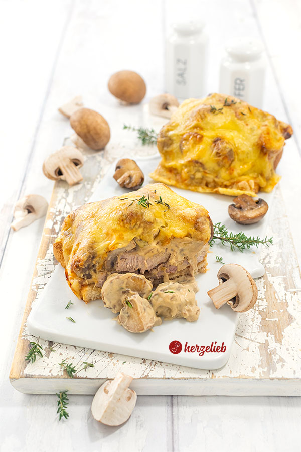Ein angeschnittener Jägerotast auf einem Porzellanbrett. Dahinter ein weiteres Toast. Dekoriert mit Thymian und Champignons