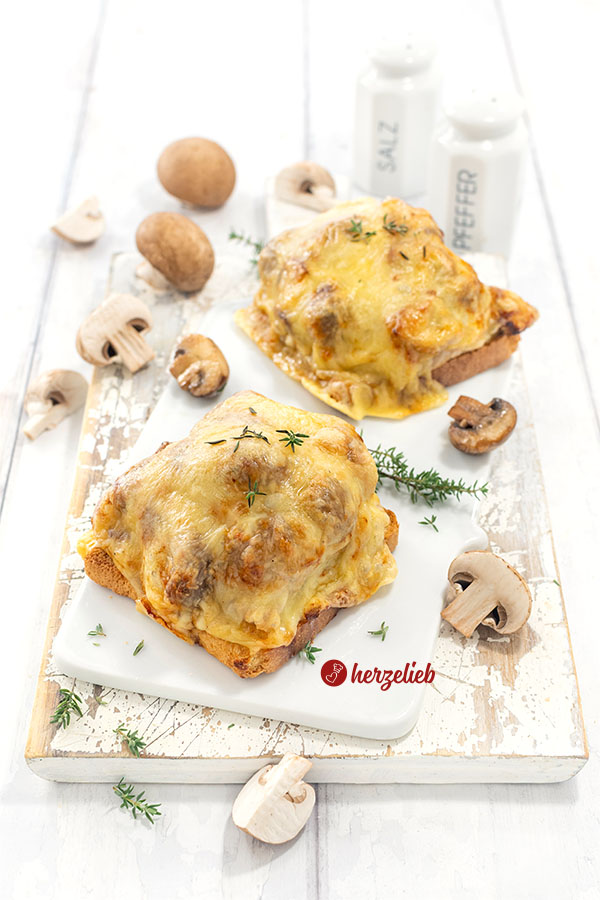 Zwei goldbaun überbackene Jägertoast auf einem Porzellanbrett. Dekoriert mit Thymian und braunen Champignons.