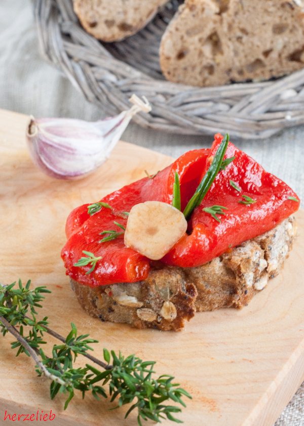 Dieses Bild zum Antipasti Rezept zeigt gegrillte Paprika auf einer Scheibe Brot mit Knoblauch und Kräutern. Im Vordergrund ein Zweig Thymian, hinter der Brotscheibe eine Knoblauchzehe. Dahinter ein Korb Brot.