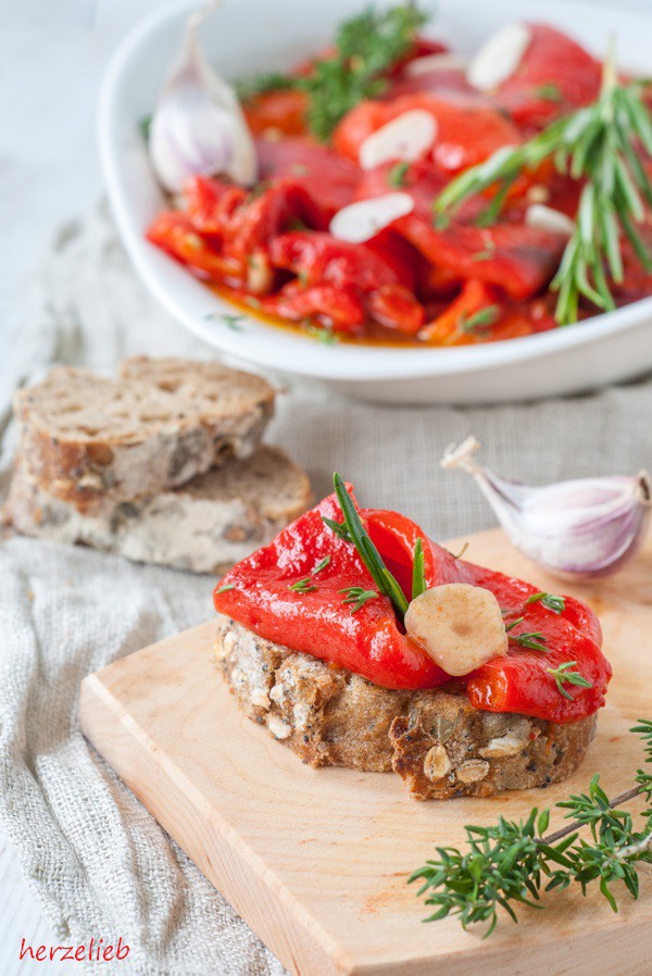 Auf diesem Bild zum Gegrillte Paprika Rezept sieht man im Vordergrund ein Brot mit einer Scheibe eingelegte Paprika, Kräutern und Knoblauch.auf einem Holzbrett. Im Vordergrund ein Zeig Thymian, hinter der Scheibe Brot eine Knoblauchzehe - Im Hintergrund eine Schale mit selbstgemachten Antipasti. Dekoriert mit Knoblauch und Kräutern.