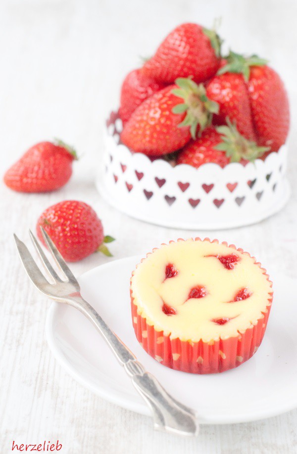 Käsekuchen-Muffins sind auch in letzter Minute schnell gebacken. Man braucht nur 30 Minuten Zeit. Ein Käsekuchenmuffin mit Erdbeerherzen auf einem weißen Teller mit einer Kuchengabel. Im Hintergrund dekoriert mit einer Erdbeerschale und 2 Erdbeeren, die daneben lieben.