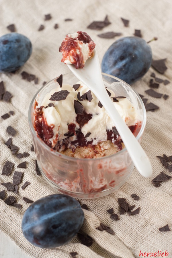Zwei Gläser mit Zwetschgenkuchen-Dessert, geschichtet aus Biskuitboden, Zwetschgenkompott und einer Mascarpone-Quark-Creme, garniert mit Schokoraspeln. Daneben liegen frische Zwetschgen und ein weißer Löffel auf einem hellen Stoffuntergrund.